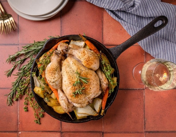 A cast iron skillet with a whole roast chicken, potatoes, and vegetables garnished with rosemary at a pairing food and pairing wine event.